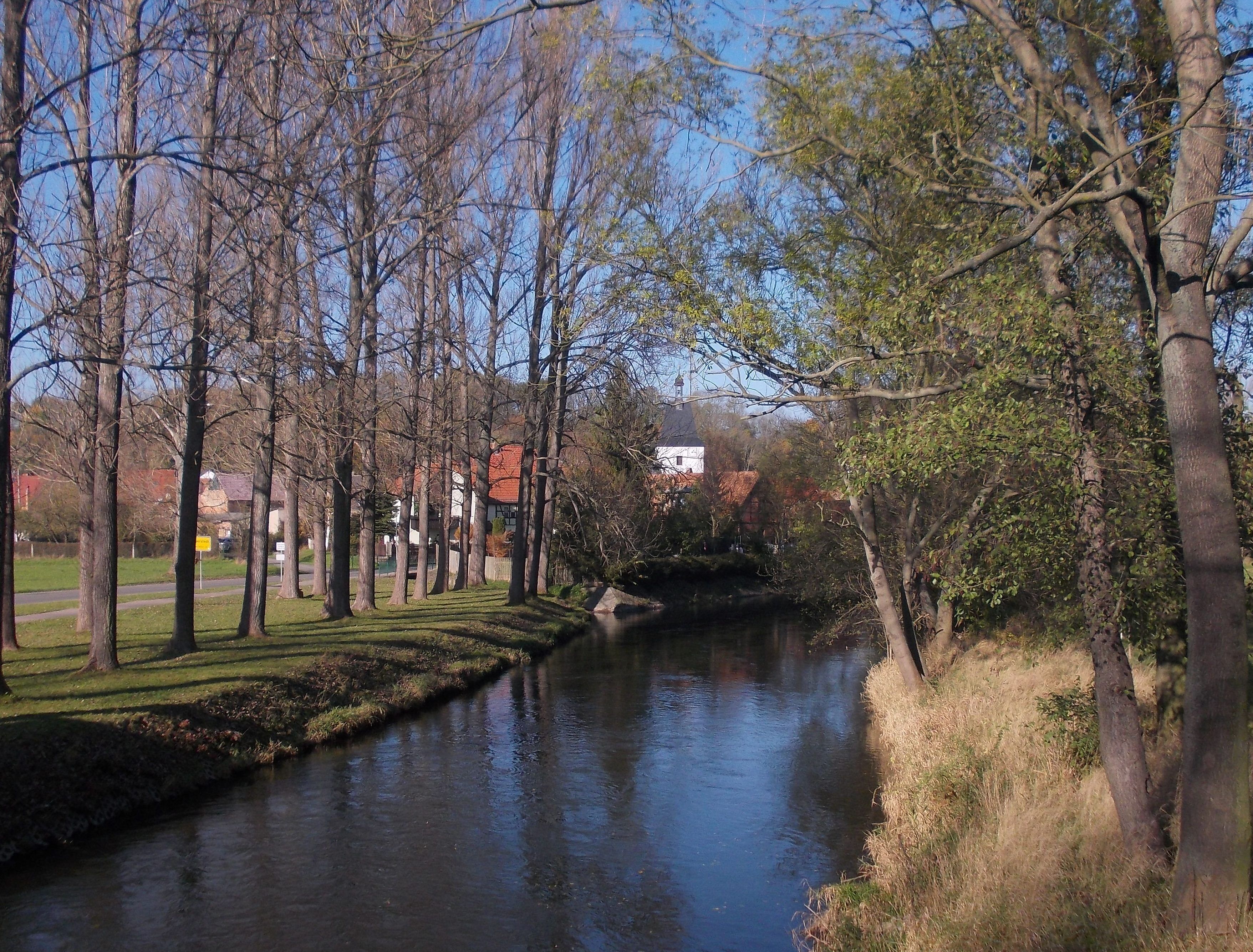 A river flanked by trees and a building with a red roof in the background.