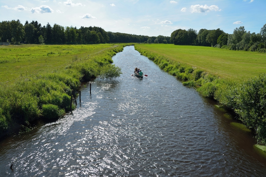 A narrow river with a boat carrying two people, surrounded by lush green fields and trees.