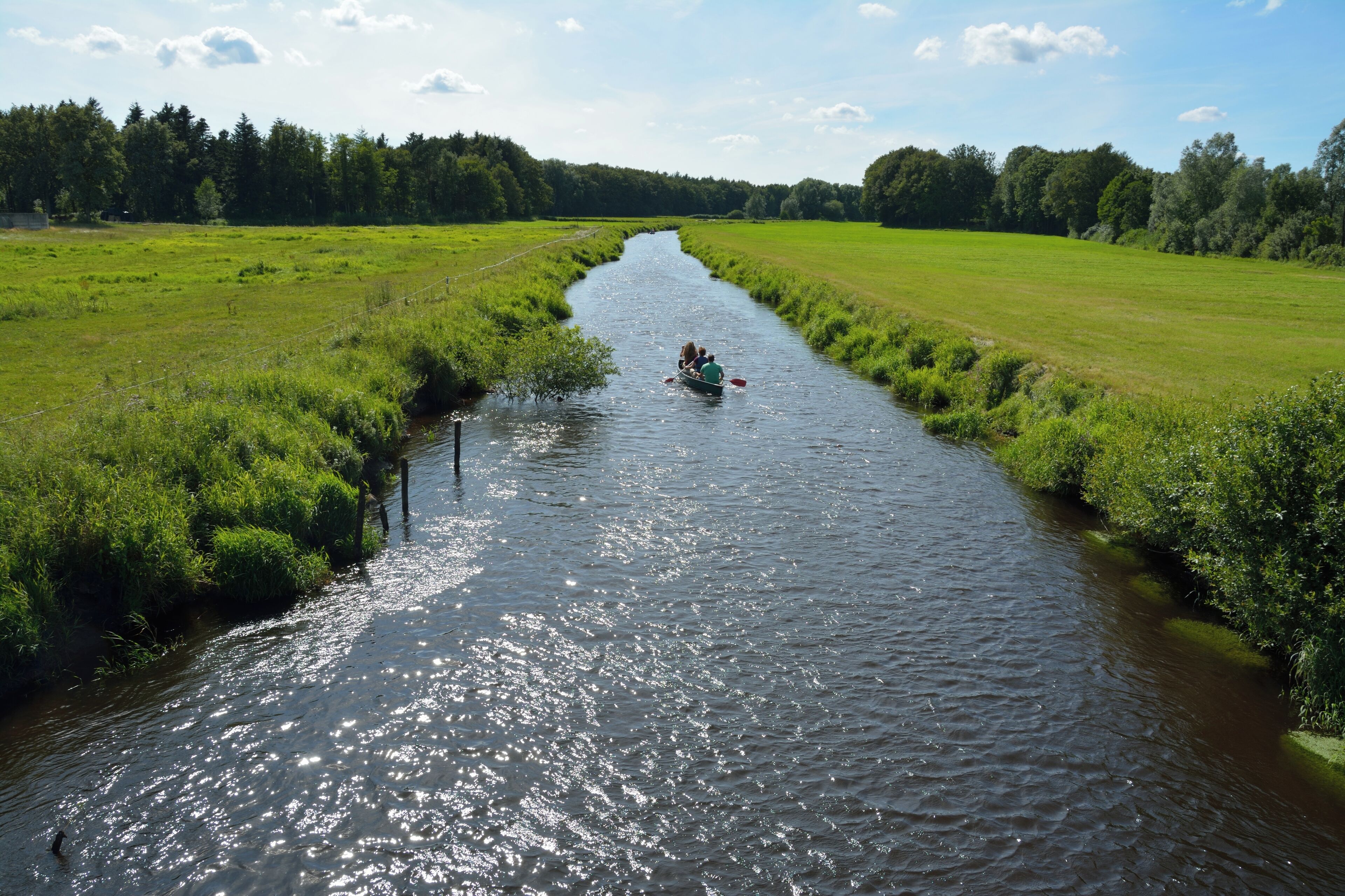 A narrow river with a boat carrying two people, surrounded by lush green fields and trees.