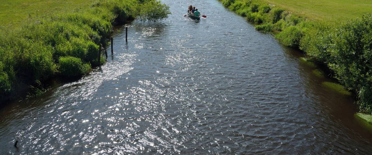 A narrow river with a boat carrying two people, surrounded by lush green fields and trees.