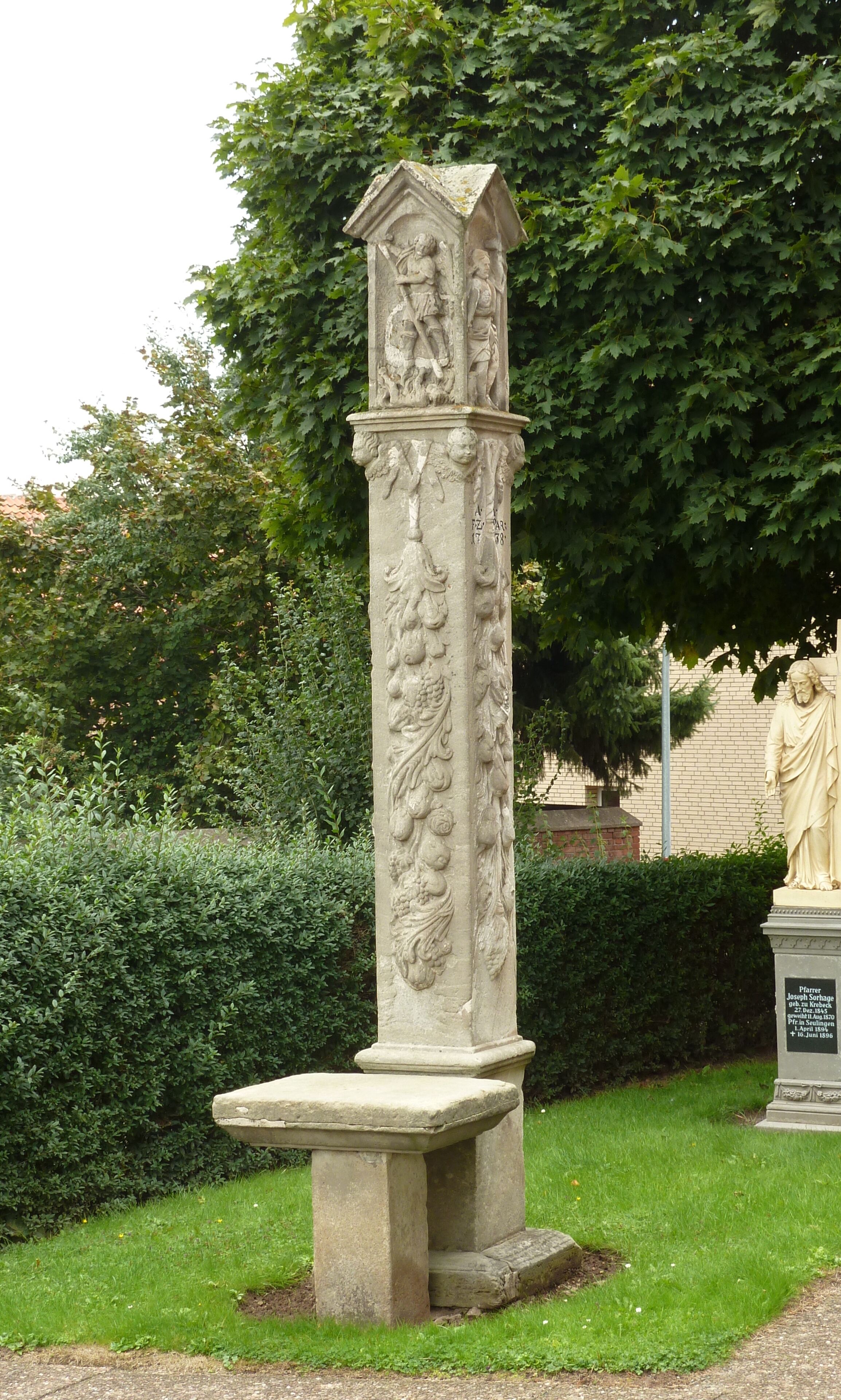 A stone monument with intricate carvings, including a figure holding a book and another figure with a staff, surrounded by lush greenery.