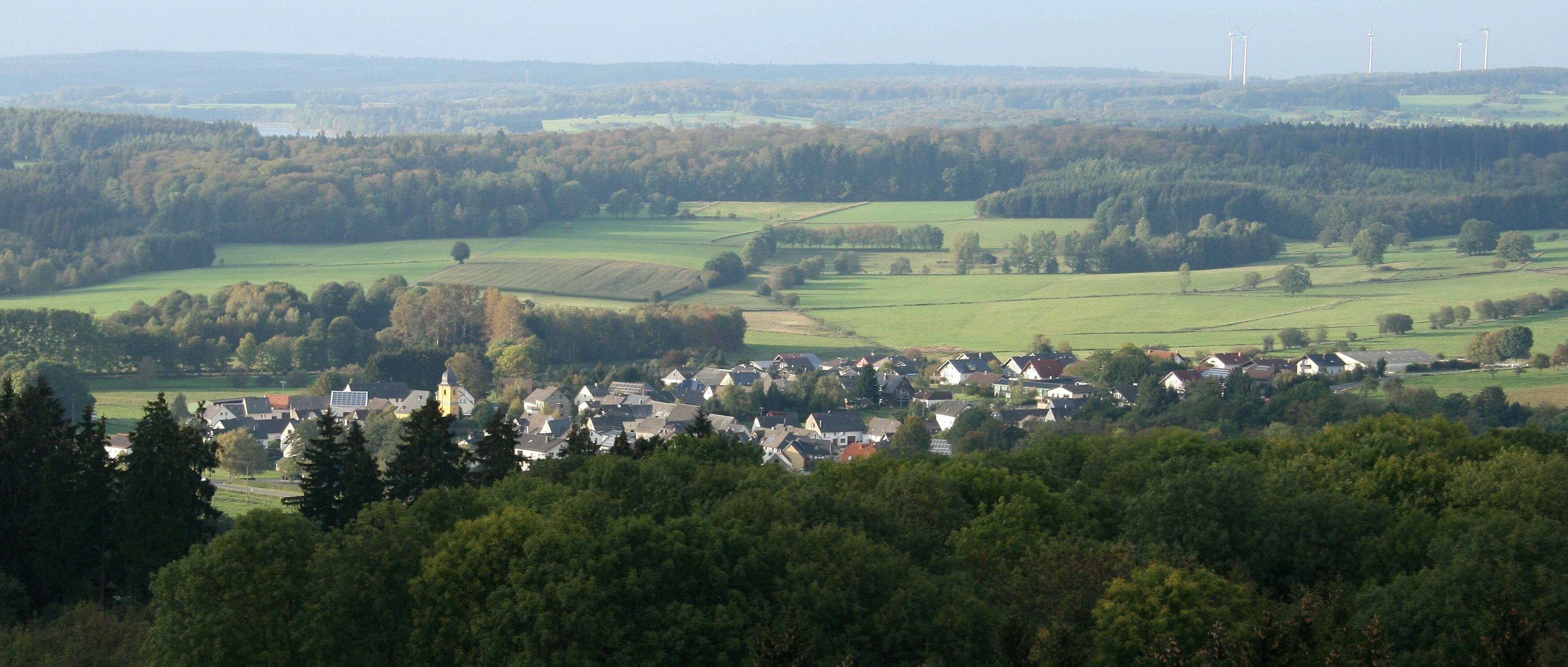 A rural landscape with a village nestled among trees and fields, with wind turbines in the distance.