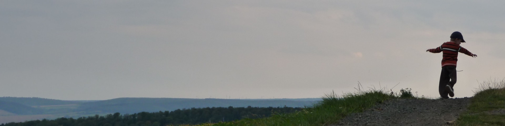 Uma pessoa em um caminho de terra com os braços estendidos, apreciando uma paisagem ondulada com campos e montanhas ao longe.