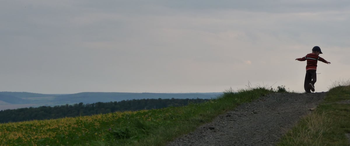 A person standing on a dirt path with arms outstretched, overlooking a hilly landscape with fields and distant mountains.