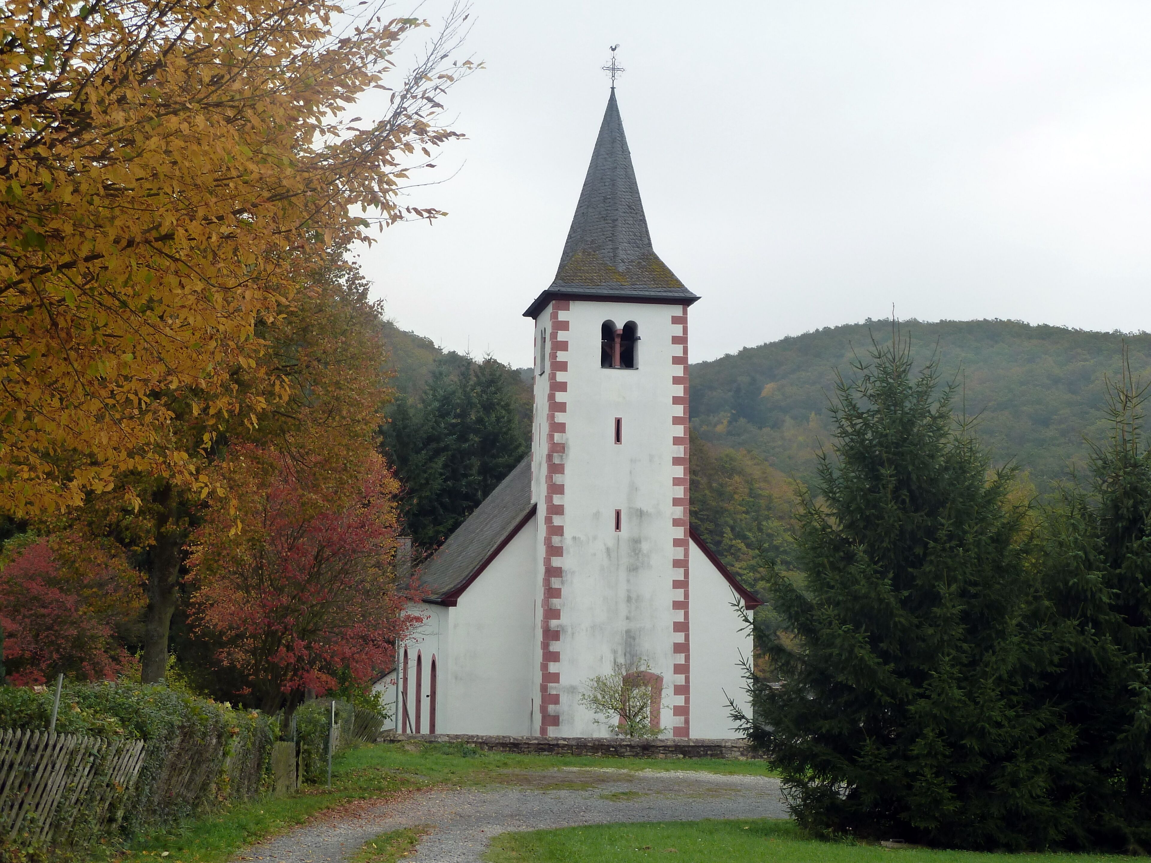 A church with a tall steeple and a weather vane, surrounded by autumn-colored trees and a hill in the background.