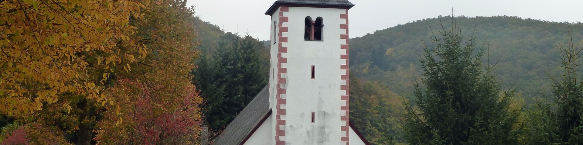 A church with a tall steeple and a weather vane, surrounded by autumn-colored trees and a hill in the background.
