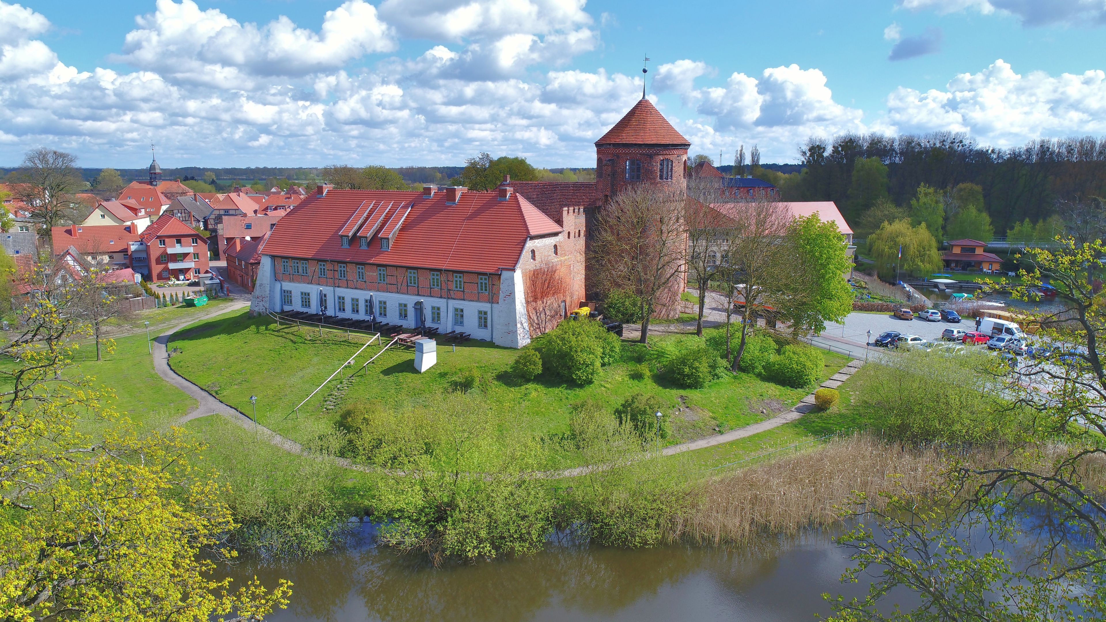 En historisk bygning med røde tegltag, et tårn og en sø i forgrunden.