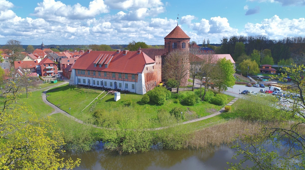 En historisk bygning med røde tegltag, et tårn og en sø i forgrunden.