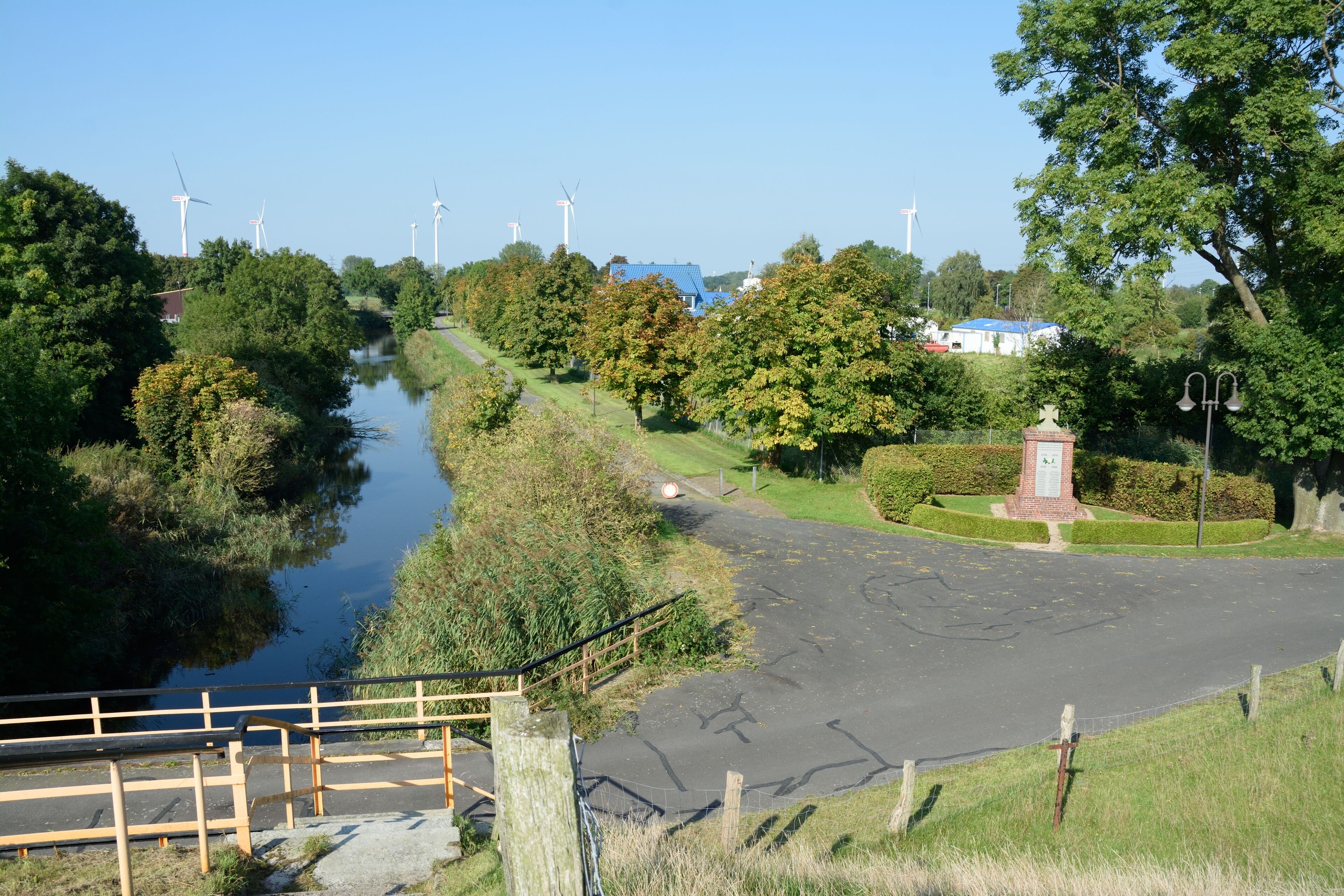 A river with wind turbines in the background, a bridge, and a park with a monument.