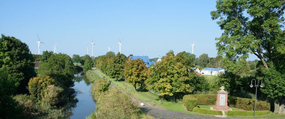 A river with wind turbines in the background, a bridge, and a park with a monument.