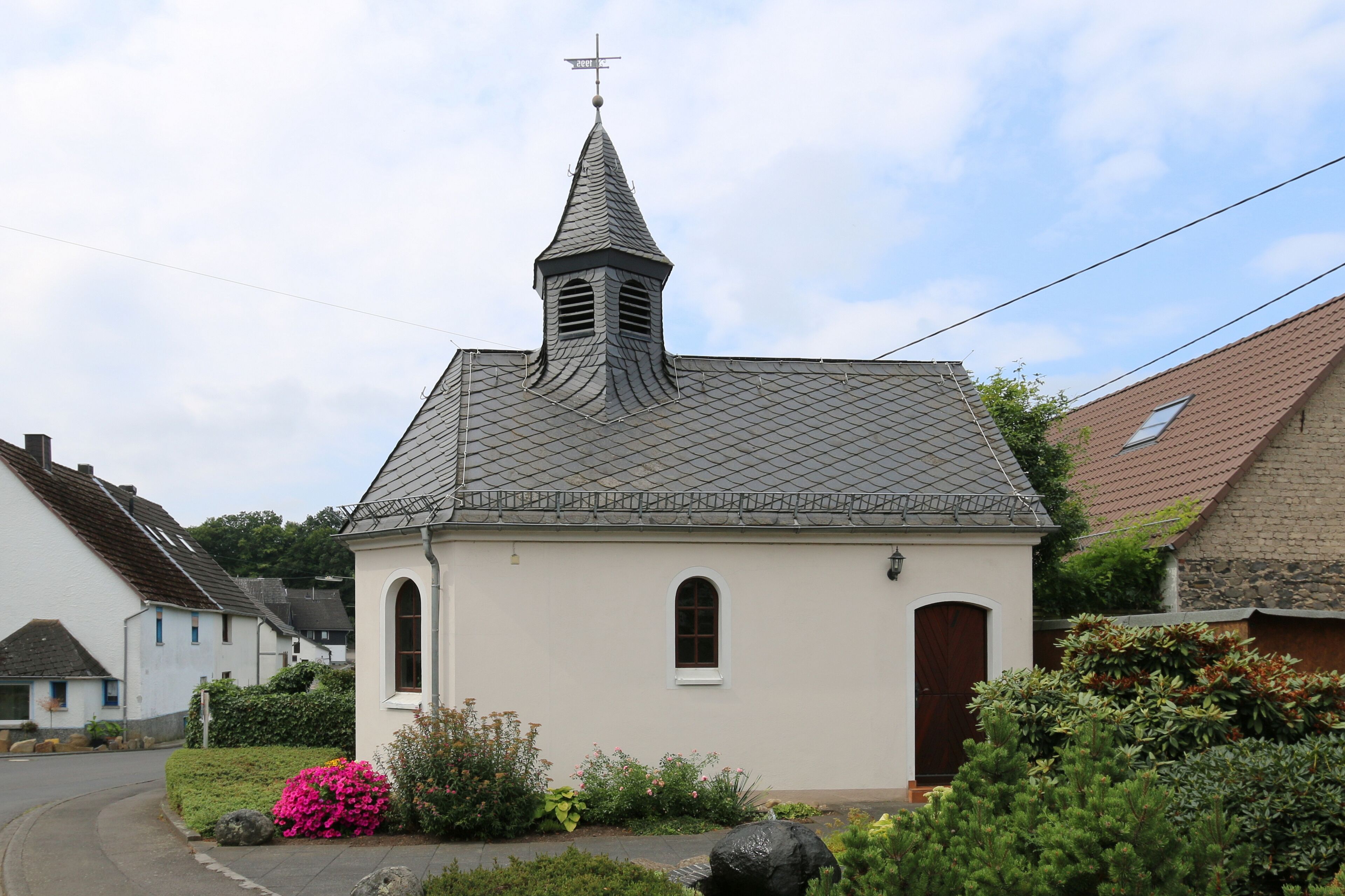 A small church with a pointed steeple and a cross on top, surrounded by greenery and a road.