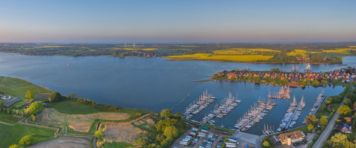 An aerial view of a harbor with numerous boats, a river, and surrounding fields.
