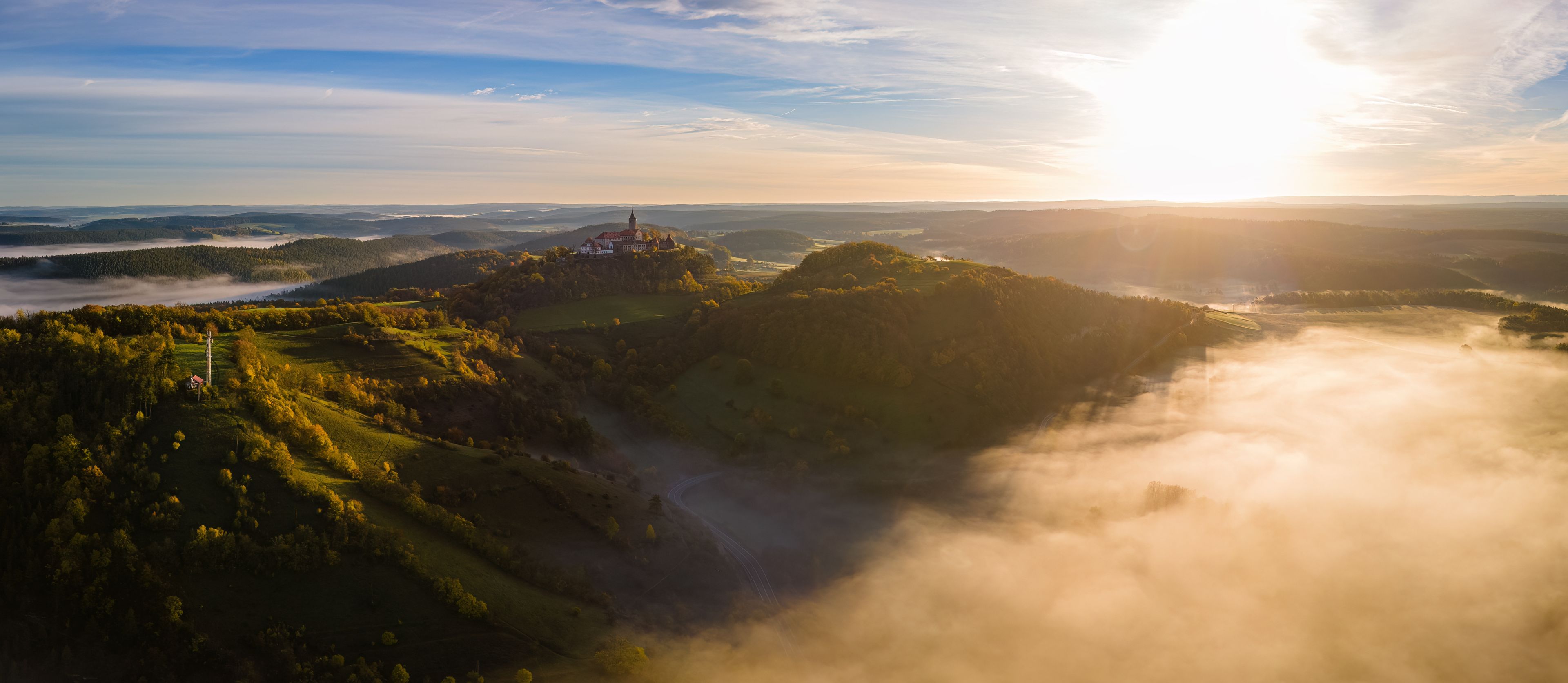 A panoramic view of a mountainous landscape with a prominent tower on a hill, surrounded by lush greenery and rolling hills.
