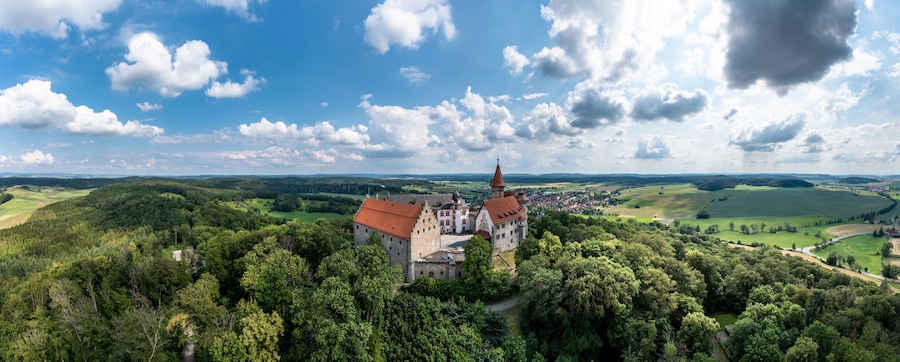 A castle surrounded by dense forest and rolling hills under a partly cloudy sky.