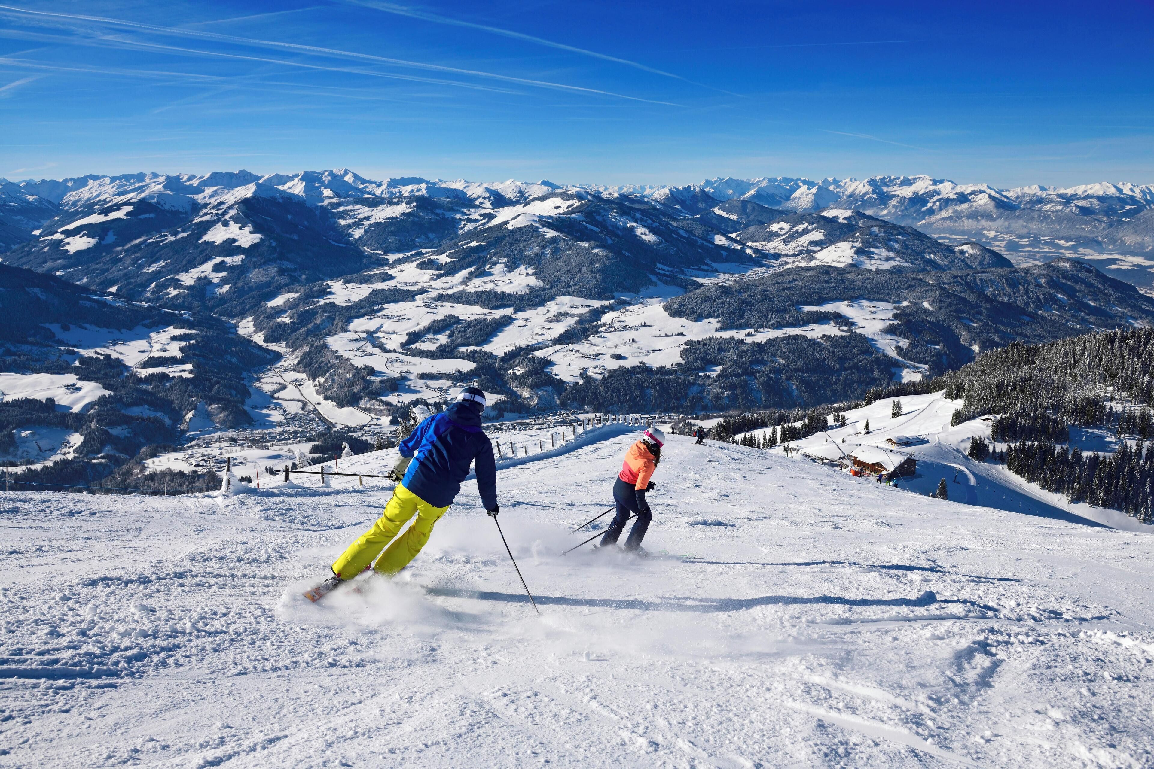Skiers on a snow-covered mountain slope with a clear blue sky and distant snow-capped peaks.