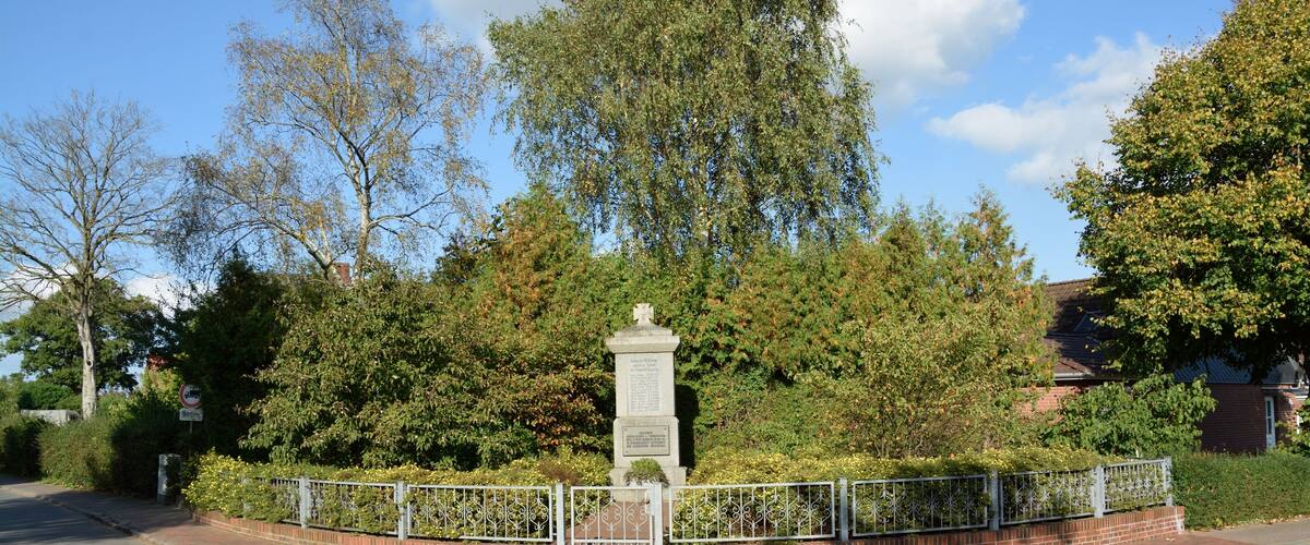 A memorial monument surrounded by a metal fence, with trees and a brick wall in the background.