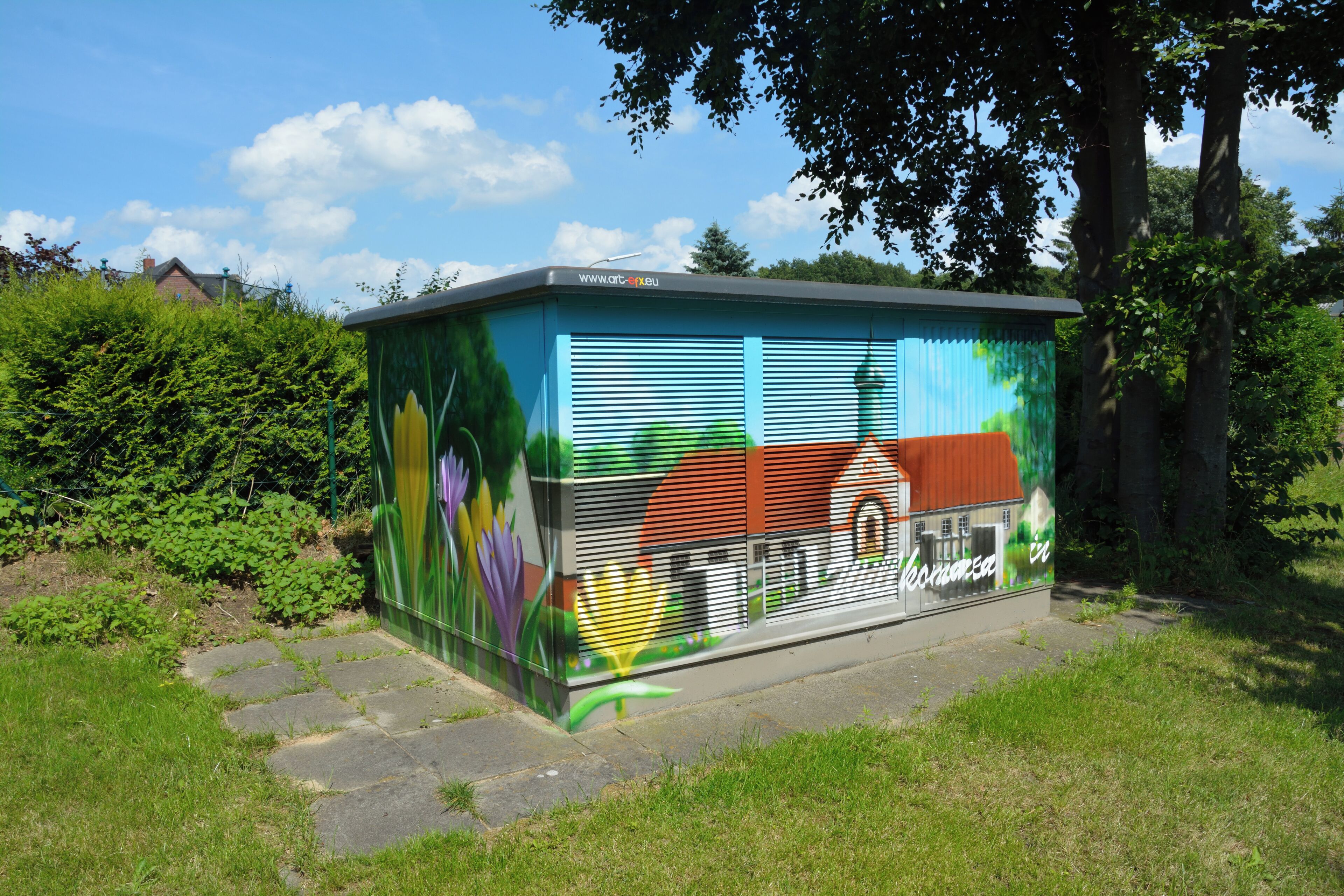 A colorful, graffiti-covered utility box in a grassy area with trees and a clear sky.