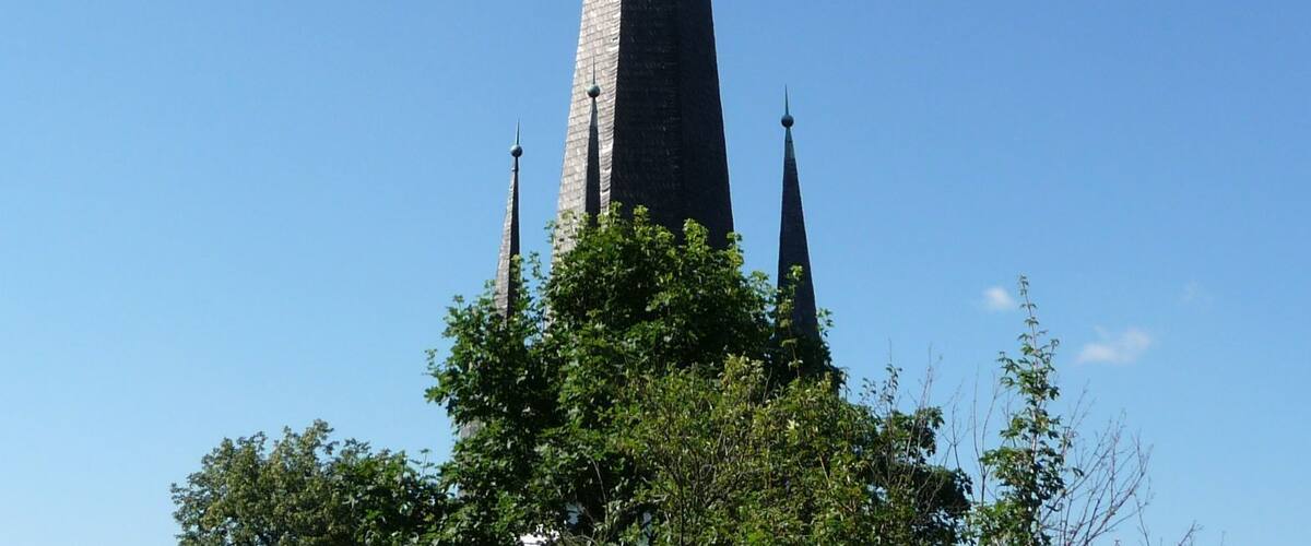 A tall church spire with three pointed tops, surrounded by trees.