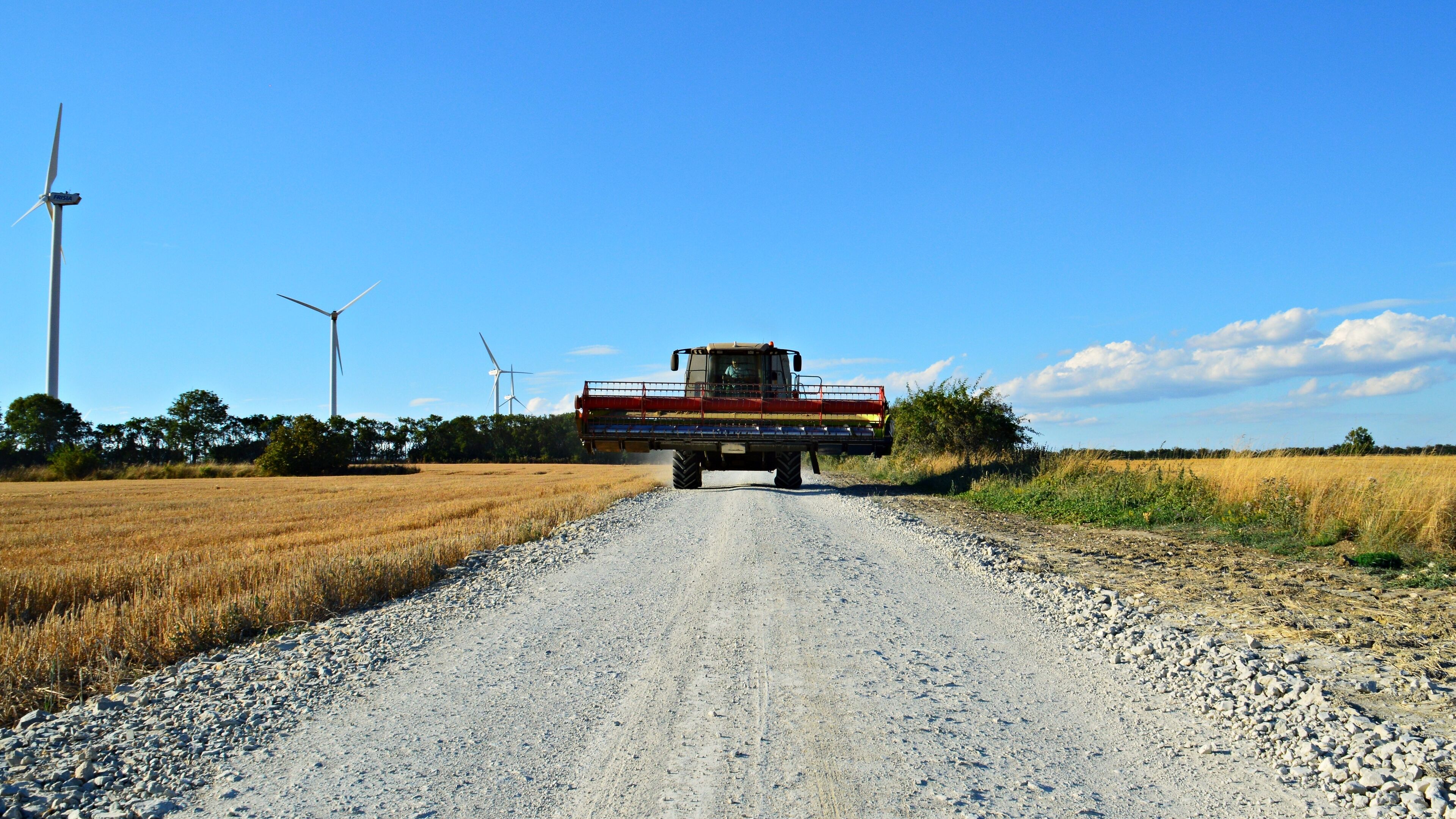 A combine harvester on a gravel road with wind turbines in the background.