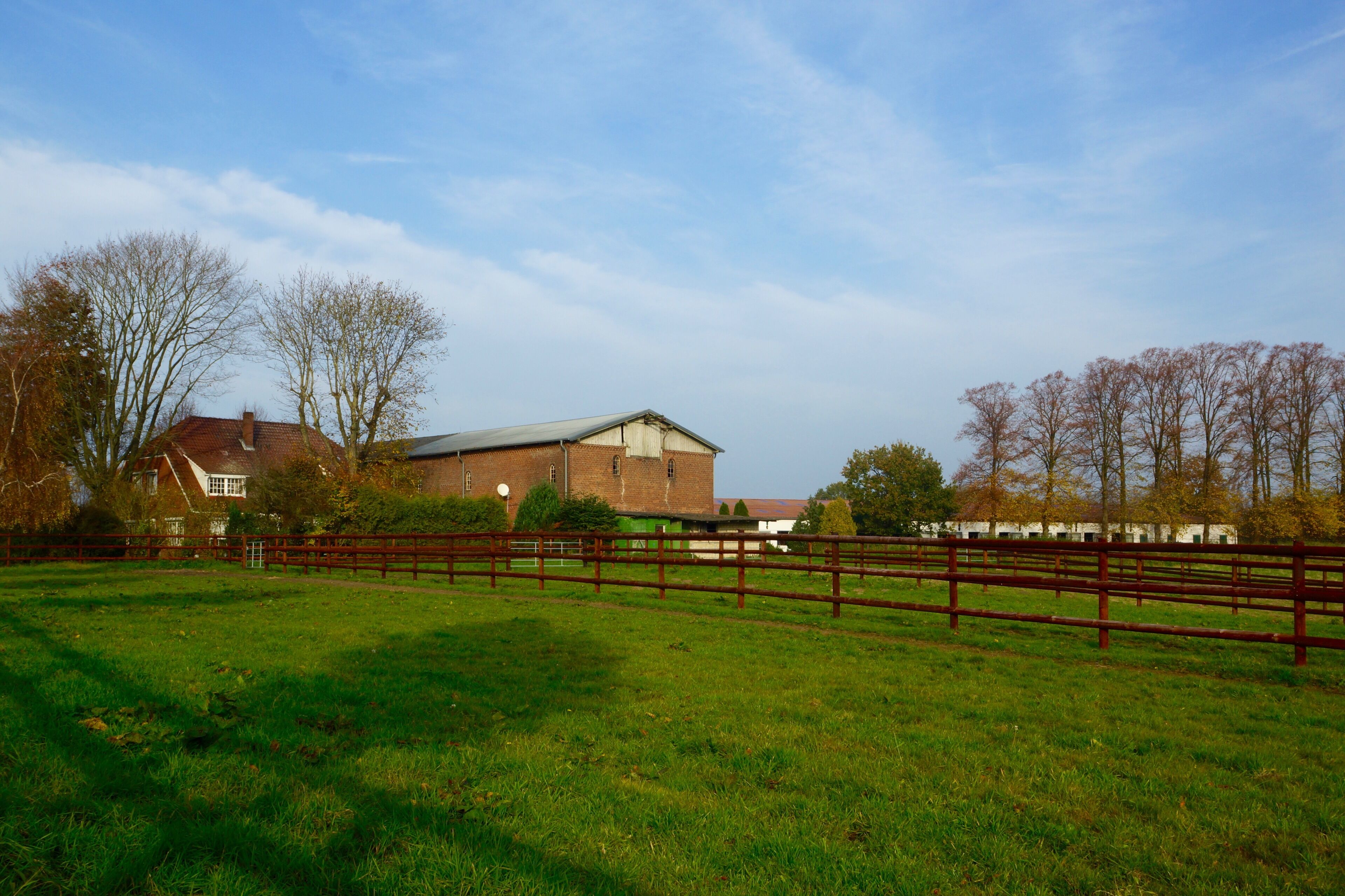 A rural landscape with a large barn, a wooden fence, and a house in the background.