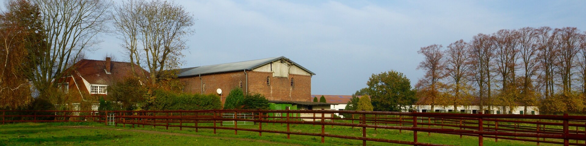 A rural landscape with a large barn, a wooden fence, and a house in the background.