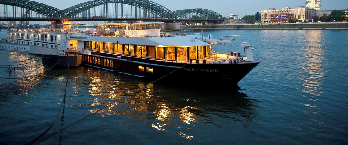 A river cruise boat named 'Serenade' docked near a bridge with a modern building in the background.