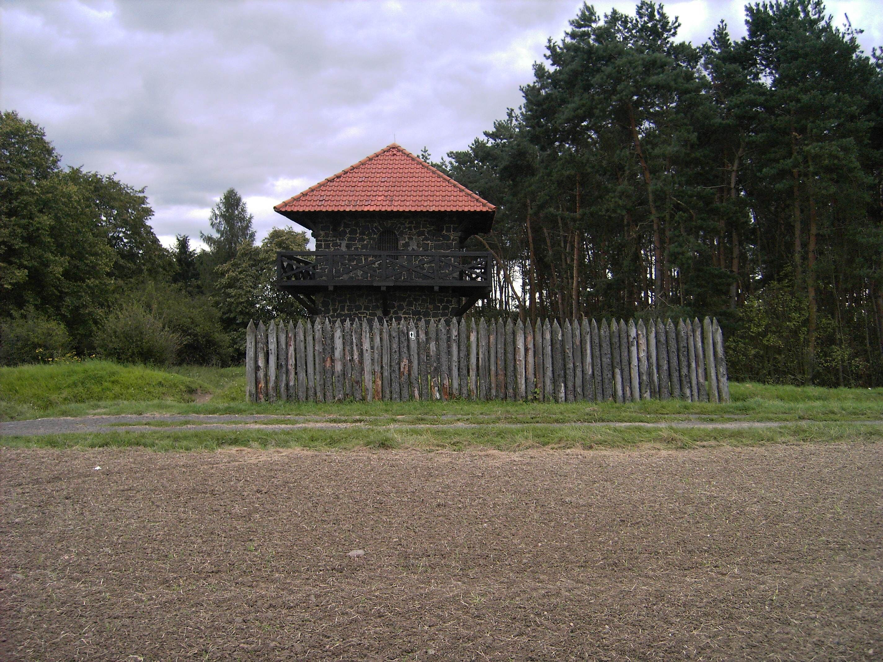A stone building with a red roof, surrounded by a wooden fence and a forest.