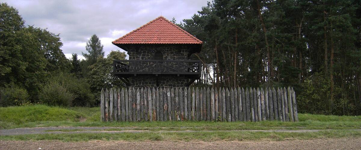 A stone building with a red roof, surrounded by a wooden fence and a forest.