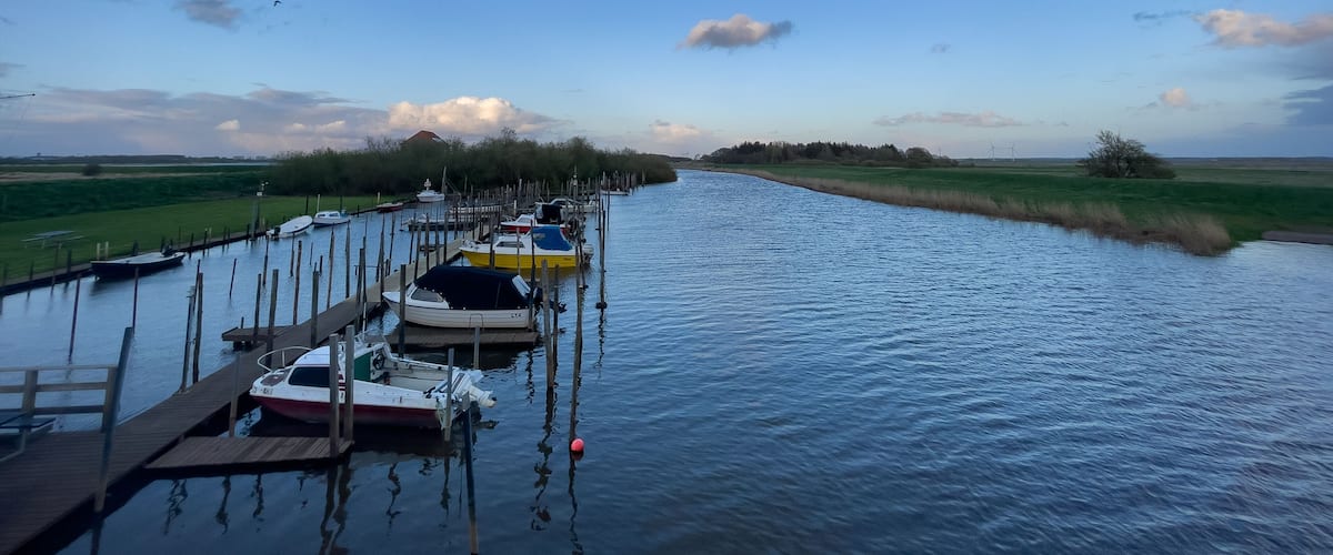 A canal with boats docked on wooden piers, surrounded by green fields and a clear sky.
