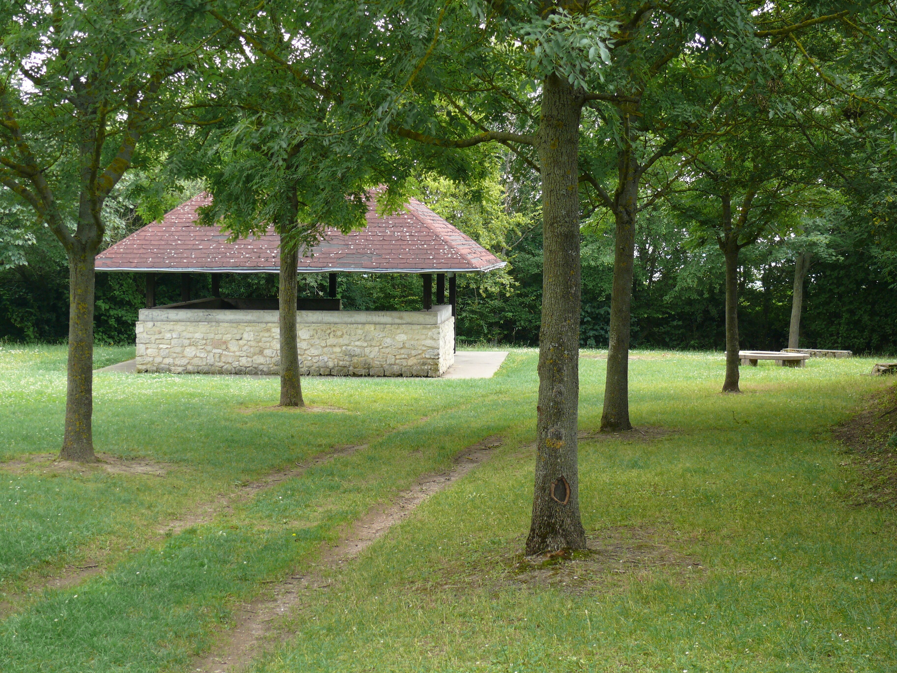 A gazebo with a stone wall and a red roof surrounded by trees and grass.