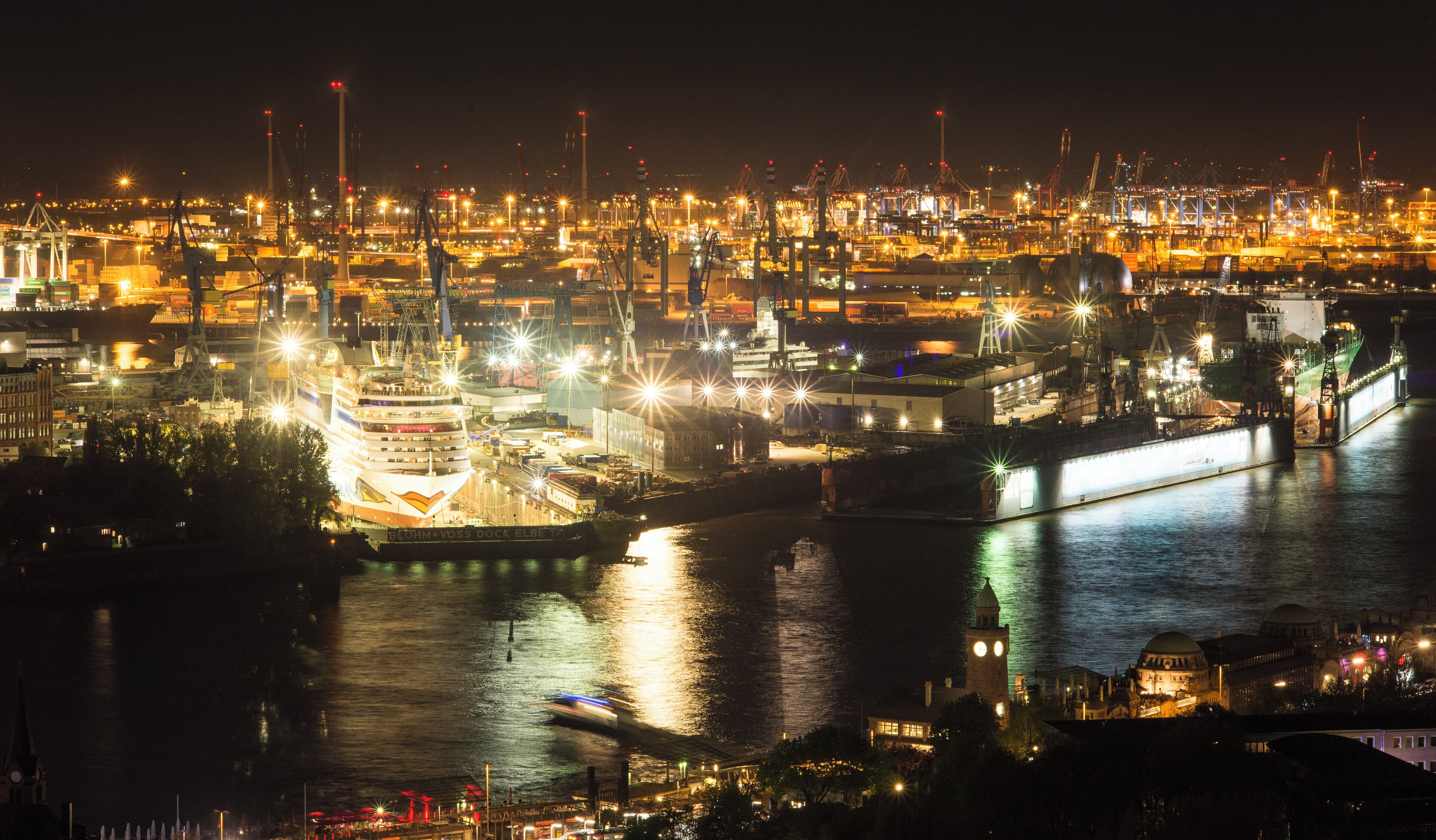 A nighttime view of a busy port with ships docked, cranes, and illuminated buildings.
