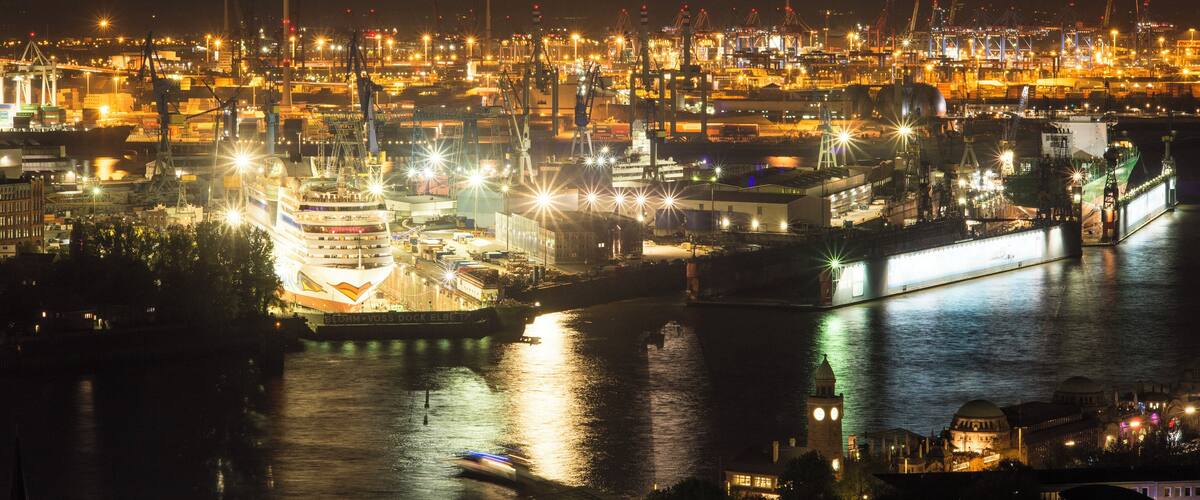 A nighttime view of a busy port with ships docked, cranes, and illuminated buildings.