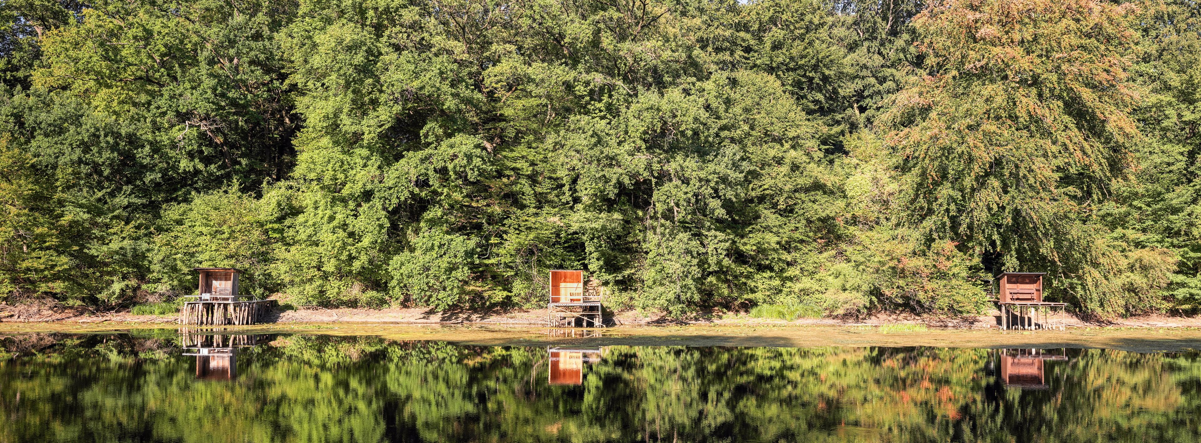 A calm lake with reflections of trees and three wooden structures on stilts along the shore.