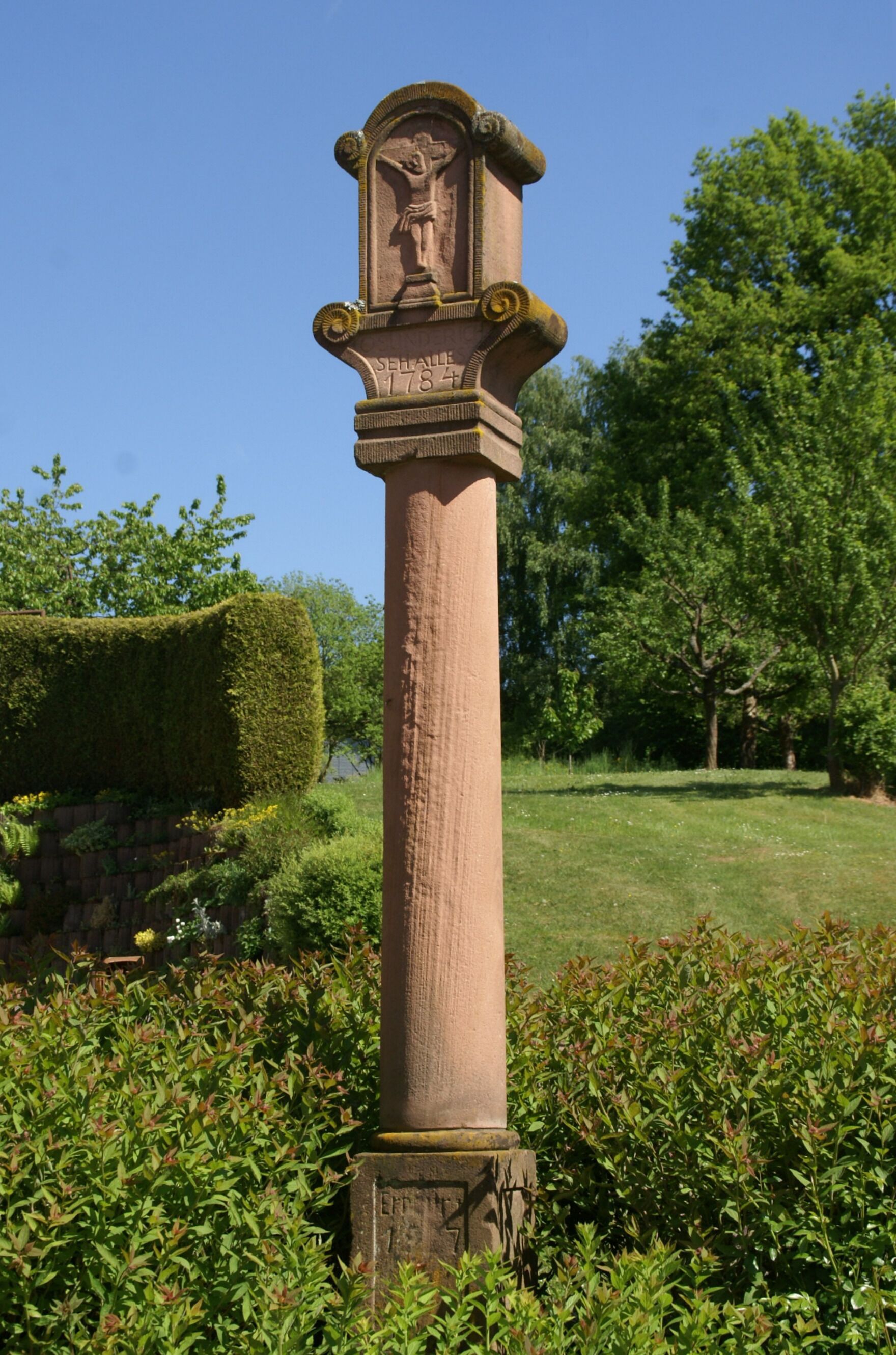 A tall stone monument with a figure, surrounded by greenery and trees.
