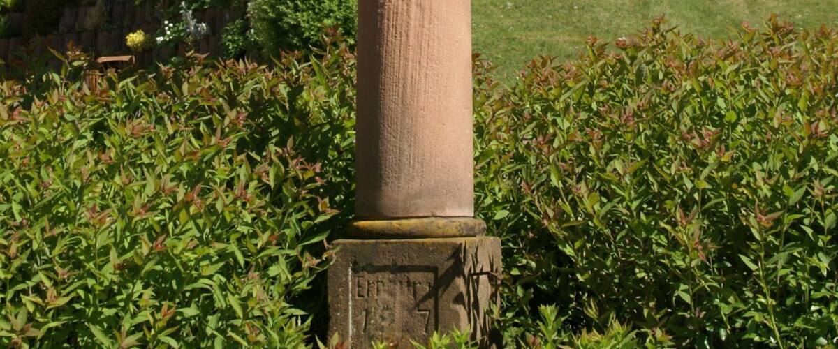 A tall stone monument with a figure, surrounded by greenery and trees.