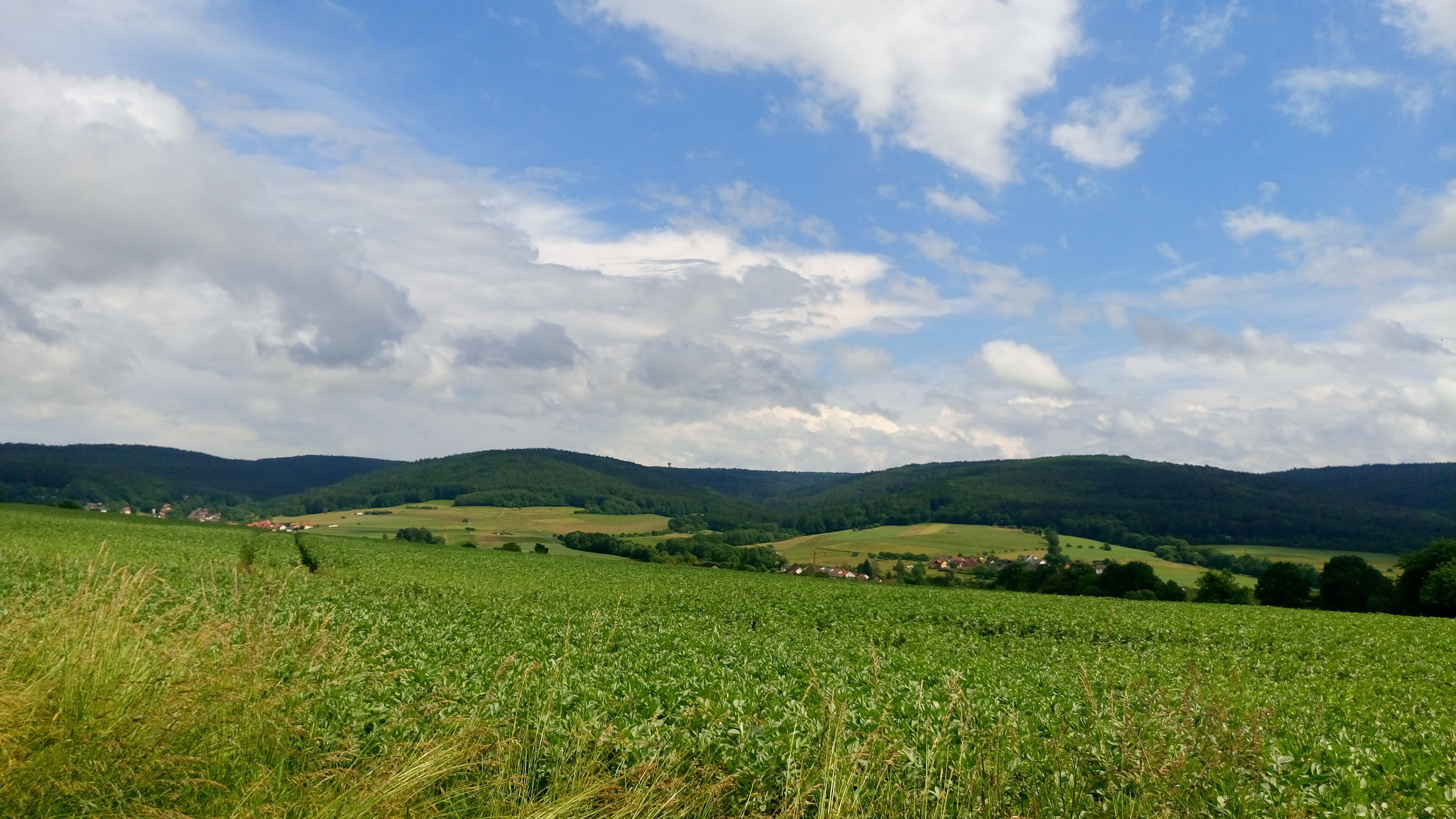 A green field with a village in the distance, surrounded by rolling hills and a partly cloudy sky.