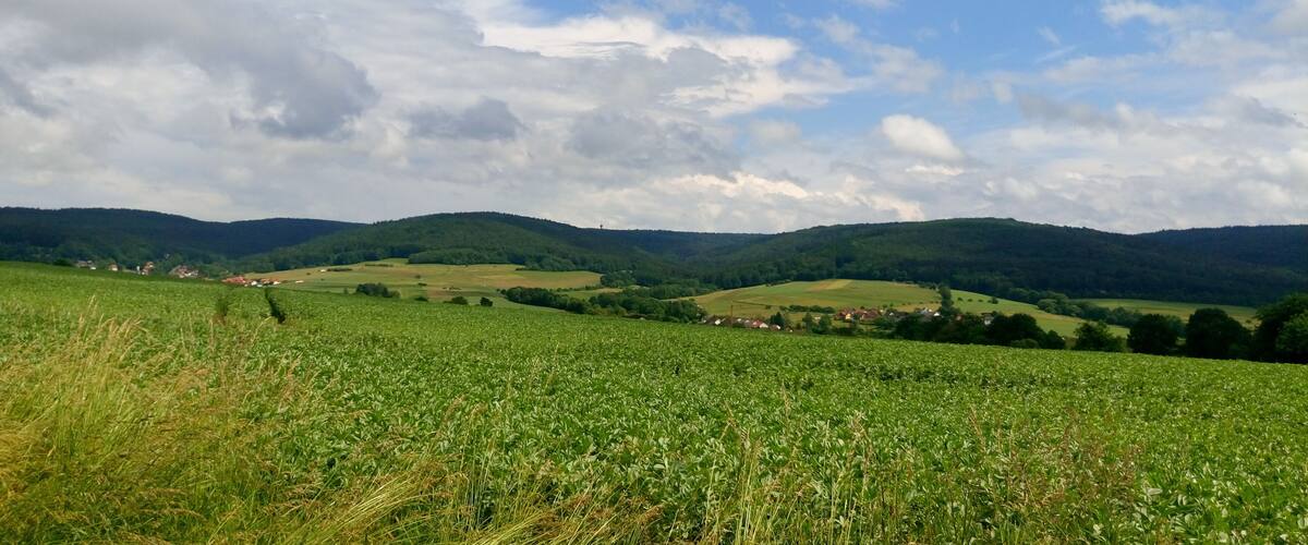 A green field with a village in the distance, surrounded by rolling hills and a partly cloudy sky.