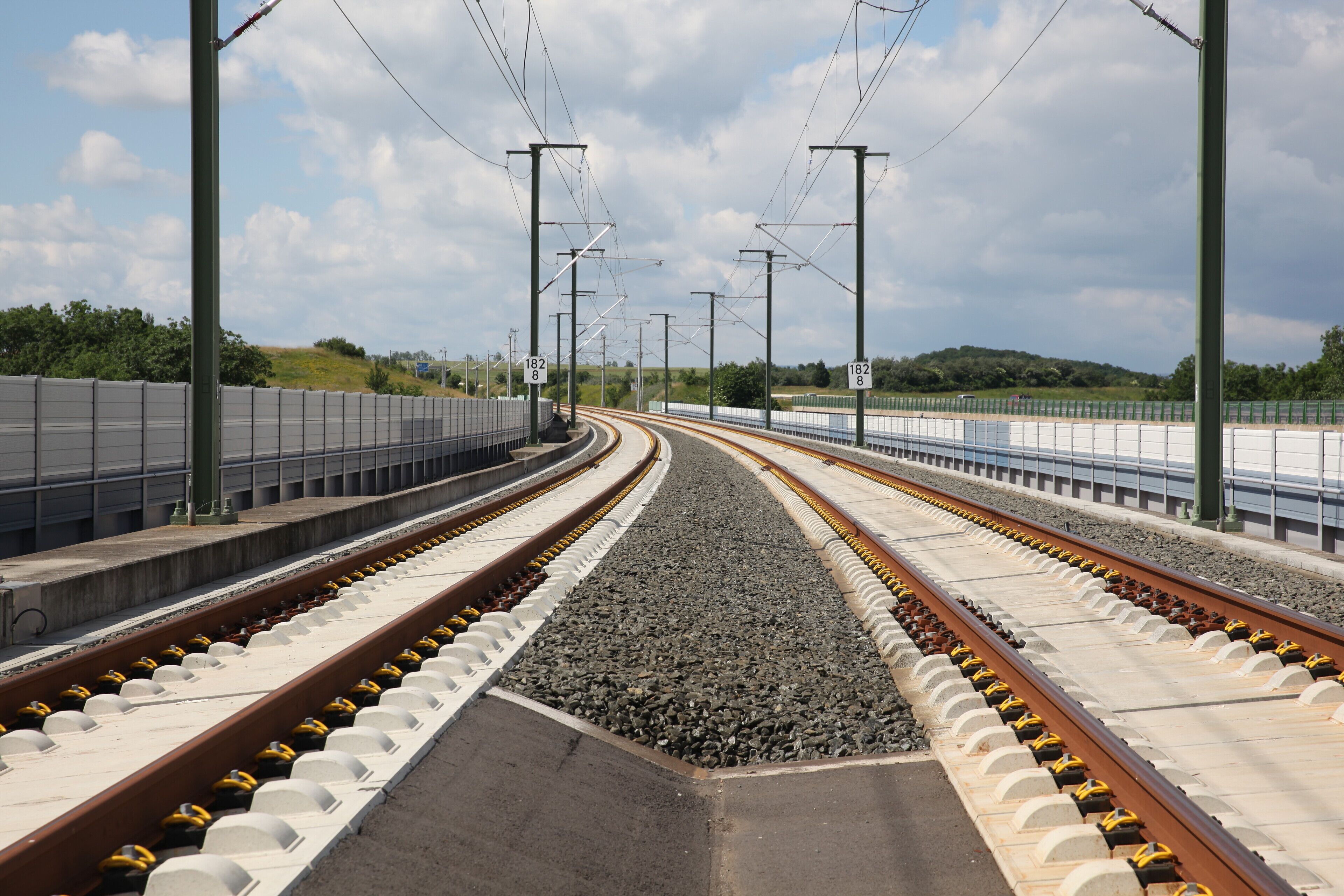 A railway track with overhead electrical lines, a concrete platform, and a grassy hill in the background.