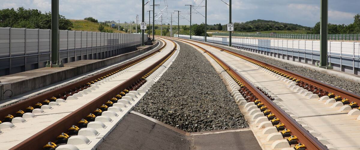 A railway track with overhead electrical lines, a concrete platform, and a grassy hill in the background.