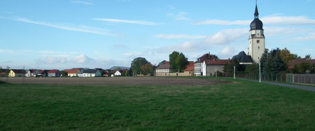 A church with a tall tower and a clock, surrounded by houses and a grassy field.