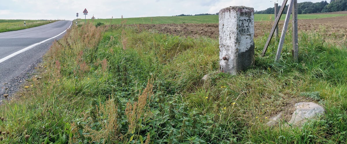 A rural road with a white post and a signboard in the foreground, surrounded by greenery and fields.