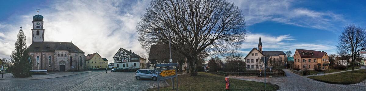 Une rue pavée bordée de bâtiments historiques, une église dotée d’un clocher et un grand arbre au centre.
