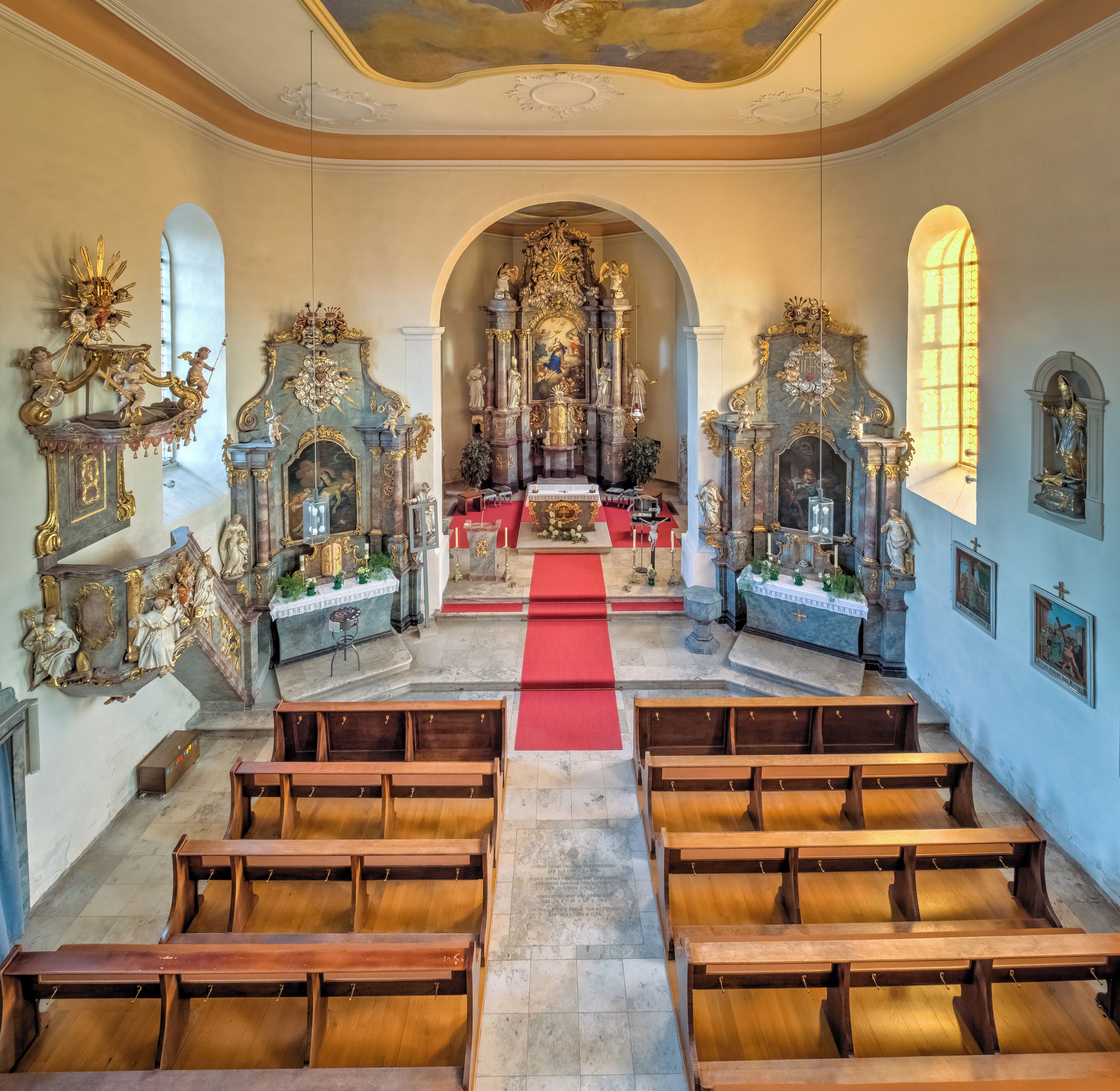 An ornate church interior with a red carpet leading to the altar, wooden pews, and elaborate religious statues and paintings.