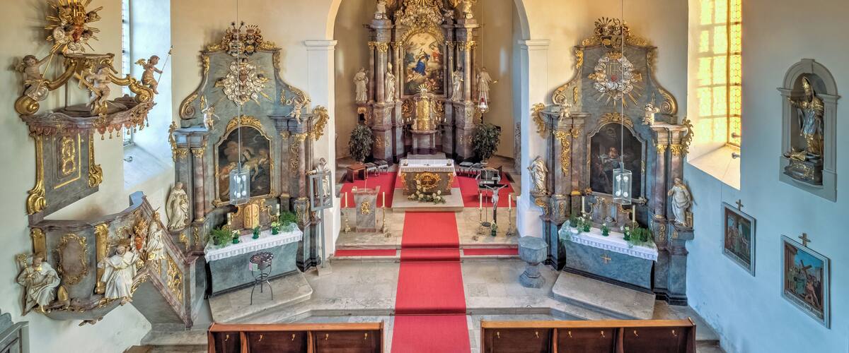 An ornate church interior with a red carpet leading to the altar, wooden pews, and elaborate religious statues and paintings.