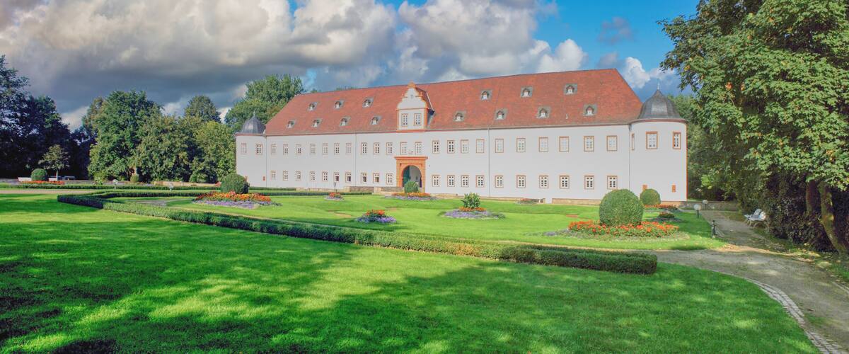 A large white building with a red roof, surrounded by a well-manicured garden with various flowers and a clear blue sky.
