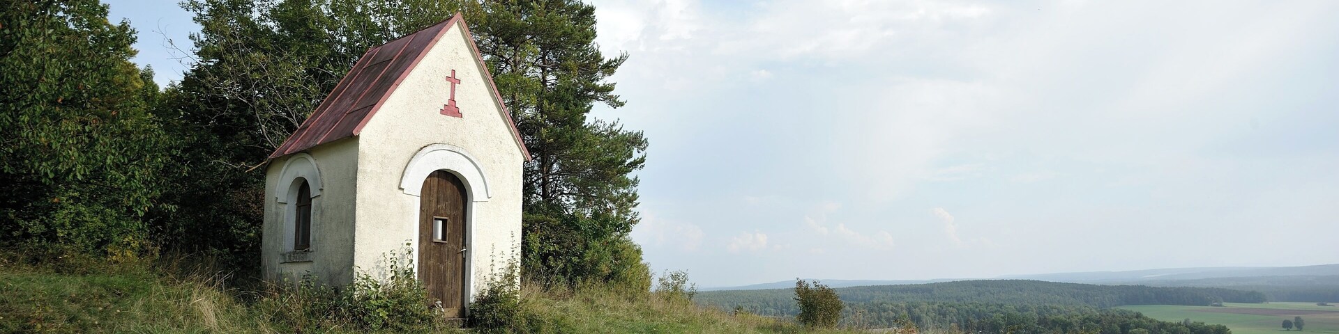 A small white church with a red cross on the roof, surrounded by green grass and trees.