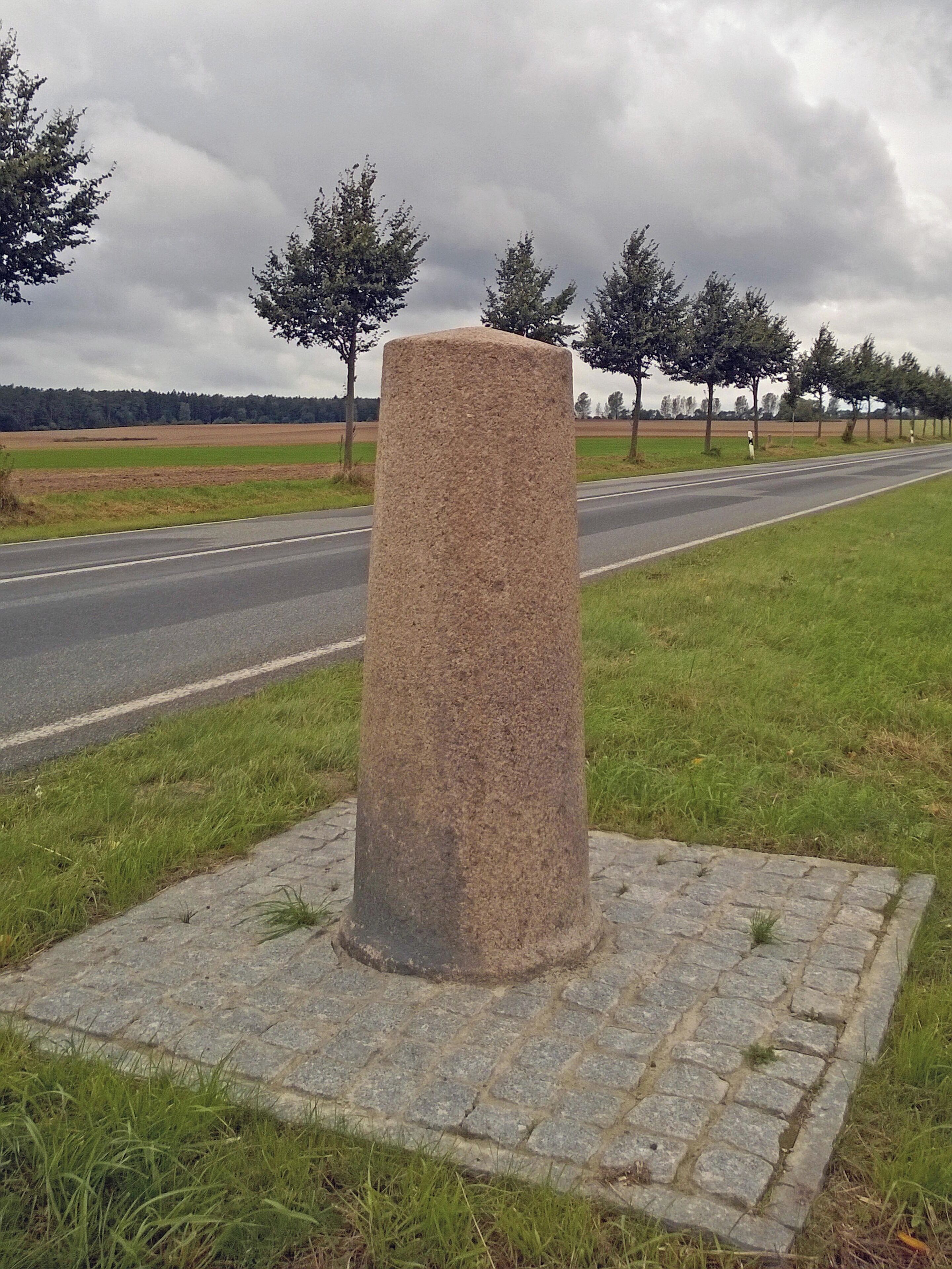 A stone monument on a paved base by a road with grass and trees in the background.