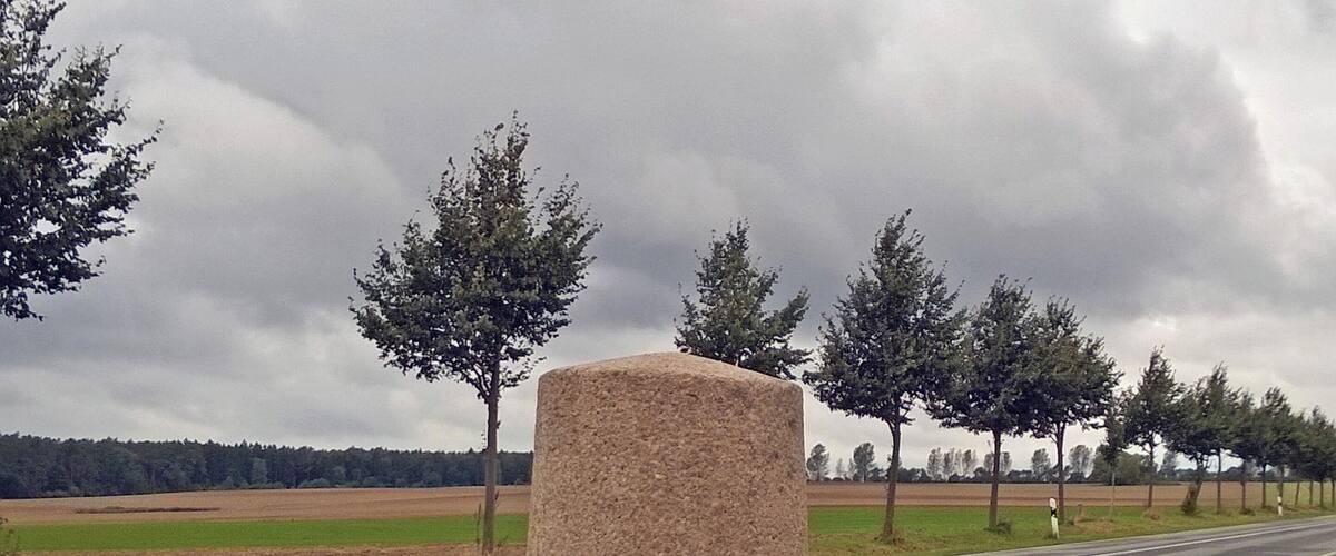 A stone monument on a paved base by a road with grass and trees in the background.
