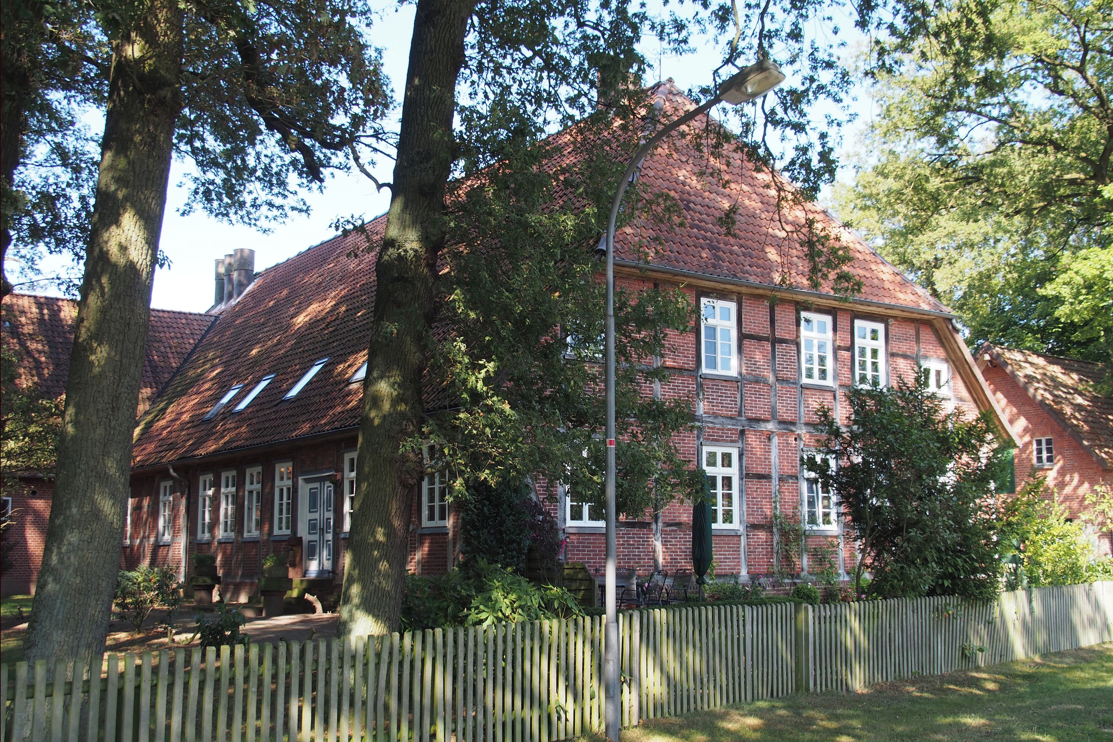 A red brick house with a sloped roof, surrounded by trees and a wooden fence.