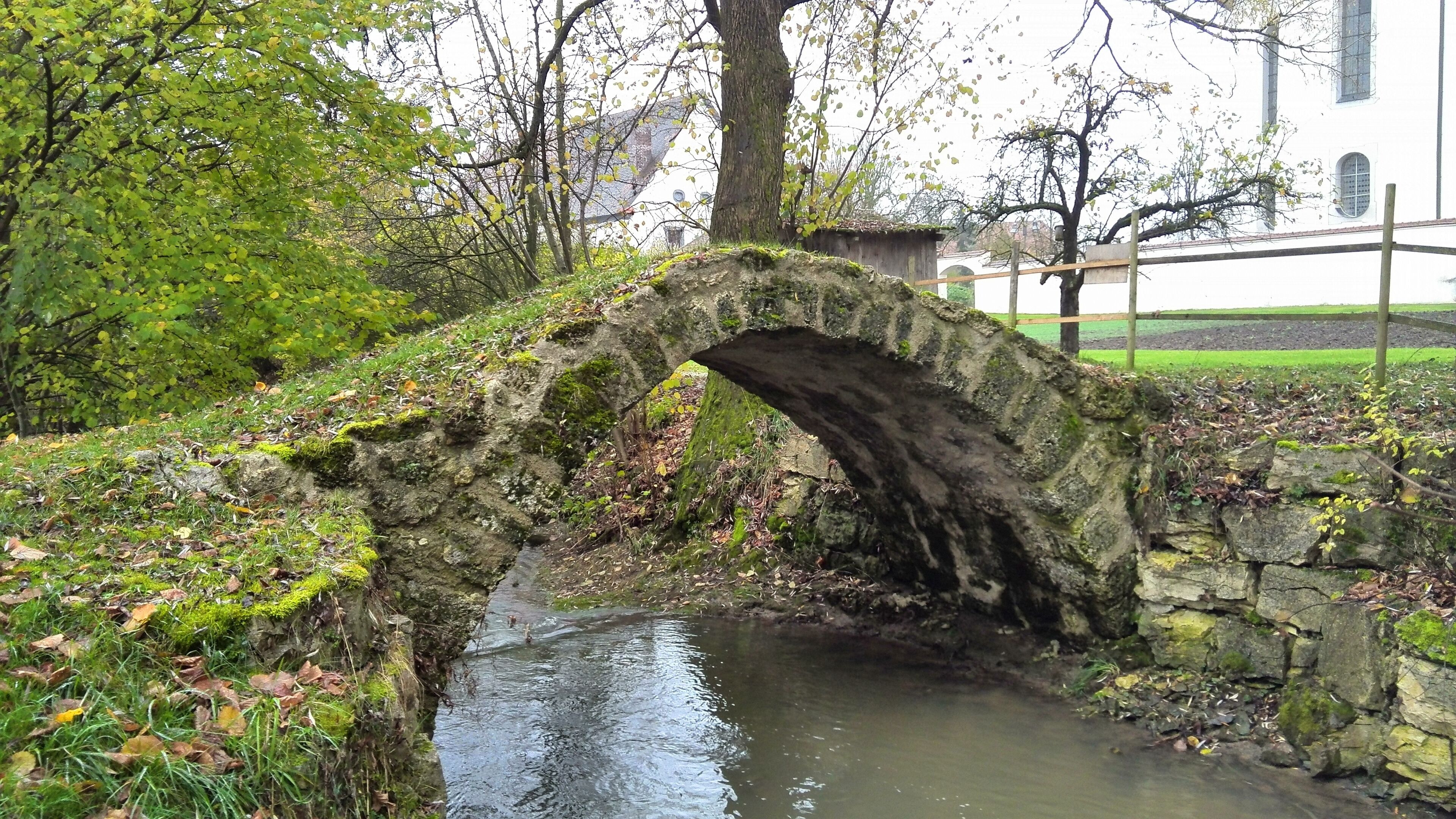 A stone bridge over a small stream in a park with trees and a building in the background.